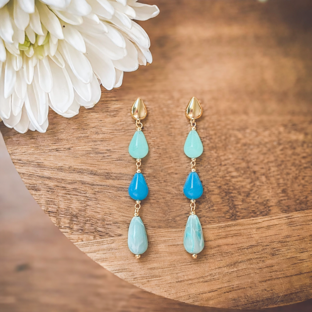 A pair of gold-toned dangle earrings featuring three teardrop-shaped stones in a gradient of light blue, medium blue, and marbled light blue, displayed on a wooden surface next to a white chrysanthemum flower