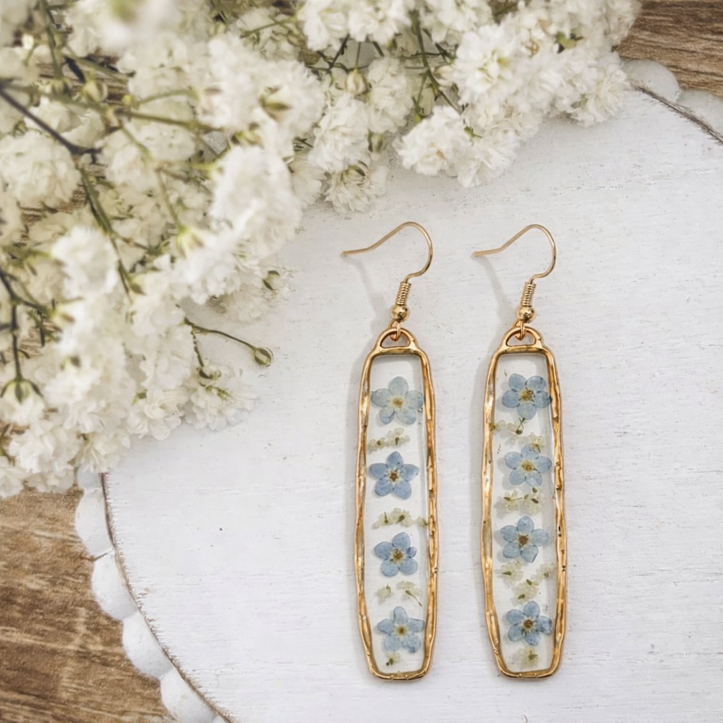 Handcrafted resin and gold-plated dangle earrings featuring delicate preserved blue forget-me-not flowers and tiny white dried blossoms, displayed on a white scalloped tray with a soft focus background of white baby's breath