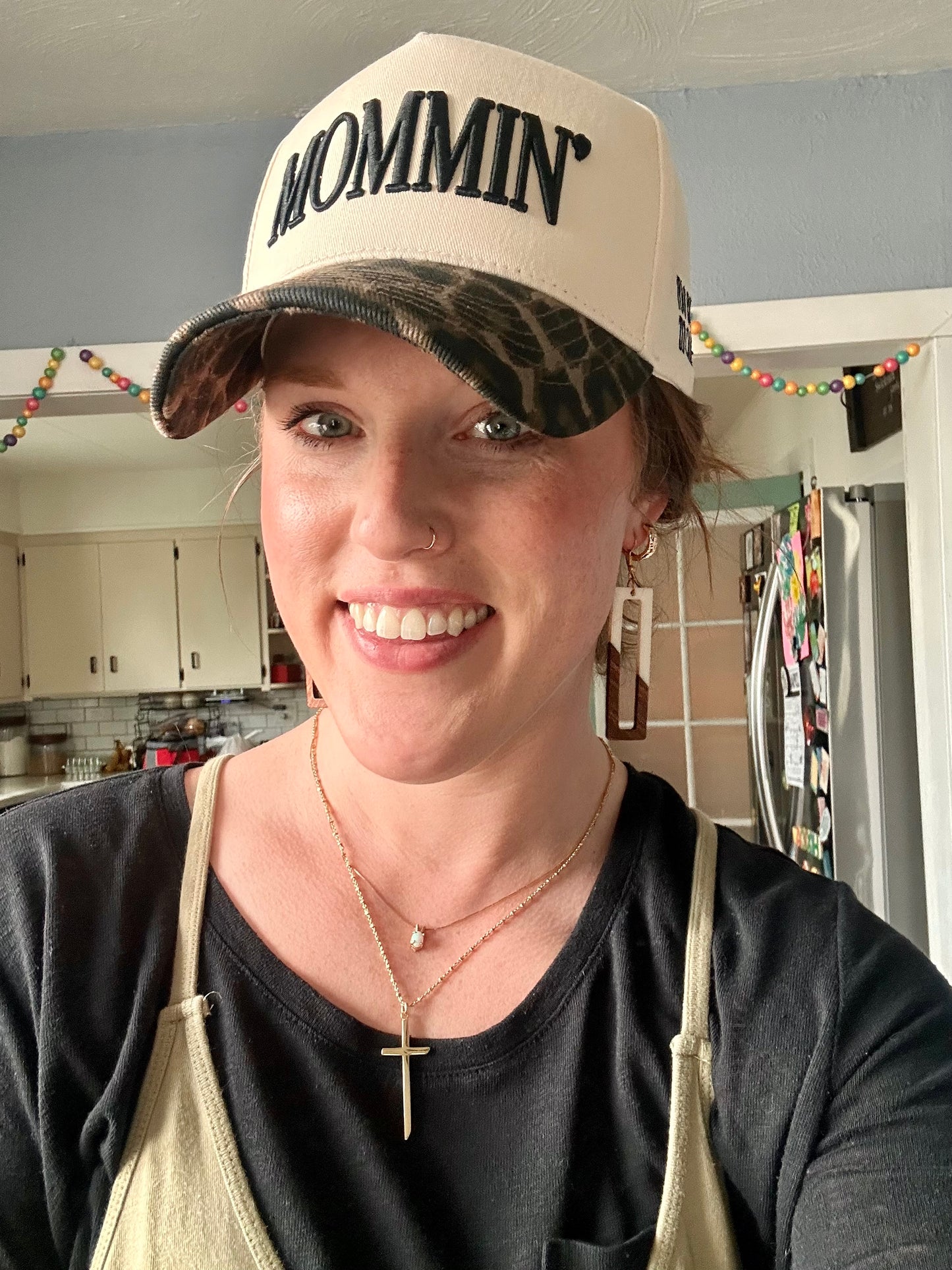 Photograph of a smiling woman in a kitchen wearing a cream "MOMMIN" hat with a camouflage brim, a nose ring, and layered gold necklaces with a cross and an opal pendant, colorful beaded garland hangs overhead