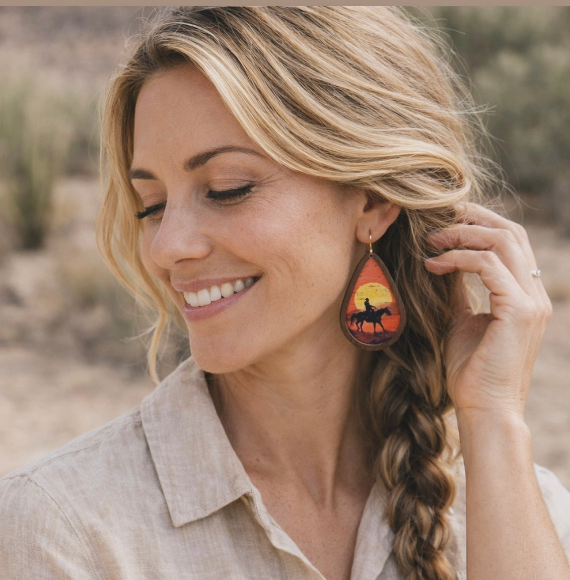 A smiling blonde woman with braided hair wearing a natural linen shirt and a teardrop-shaped wooden earring featuring a cowboy on horseback silhouette against a vibrant orange and yellow sunset in a desert landscape