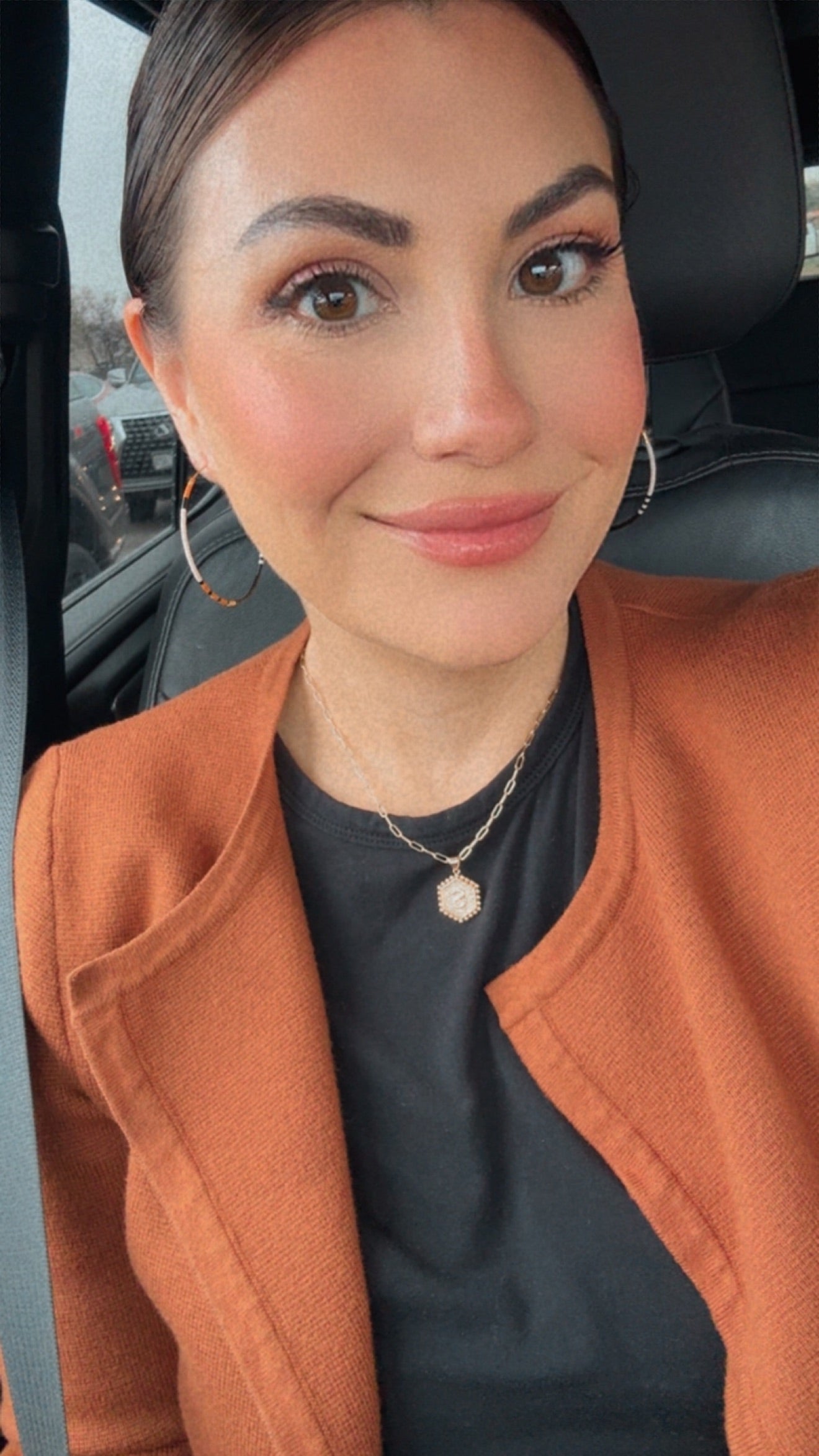 A smiling woman with rosy cheeks and dark hair pulled back wears a rust-orange open cardigan over a black top, a silver hexagonal initial pendant necklace, and white-orange-black beaded hoop earrings in a car interior