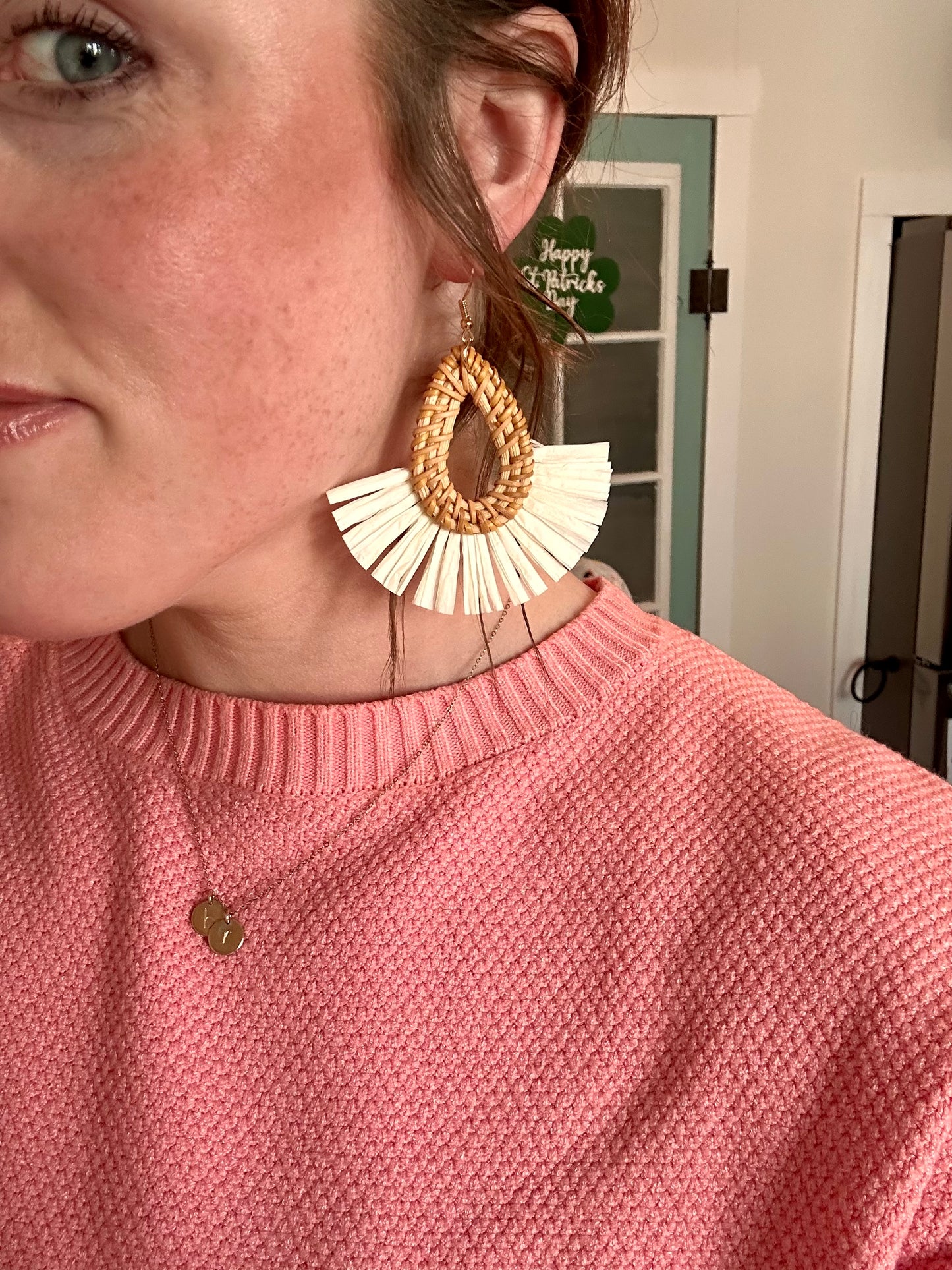 Photograph of a woman's profile featuring a large woven raffia and white fringe earring, a salmon pink chunky knit sweater, and a green Happy St Patrick's Day shamrock decoration in the background