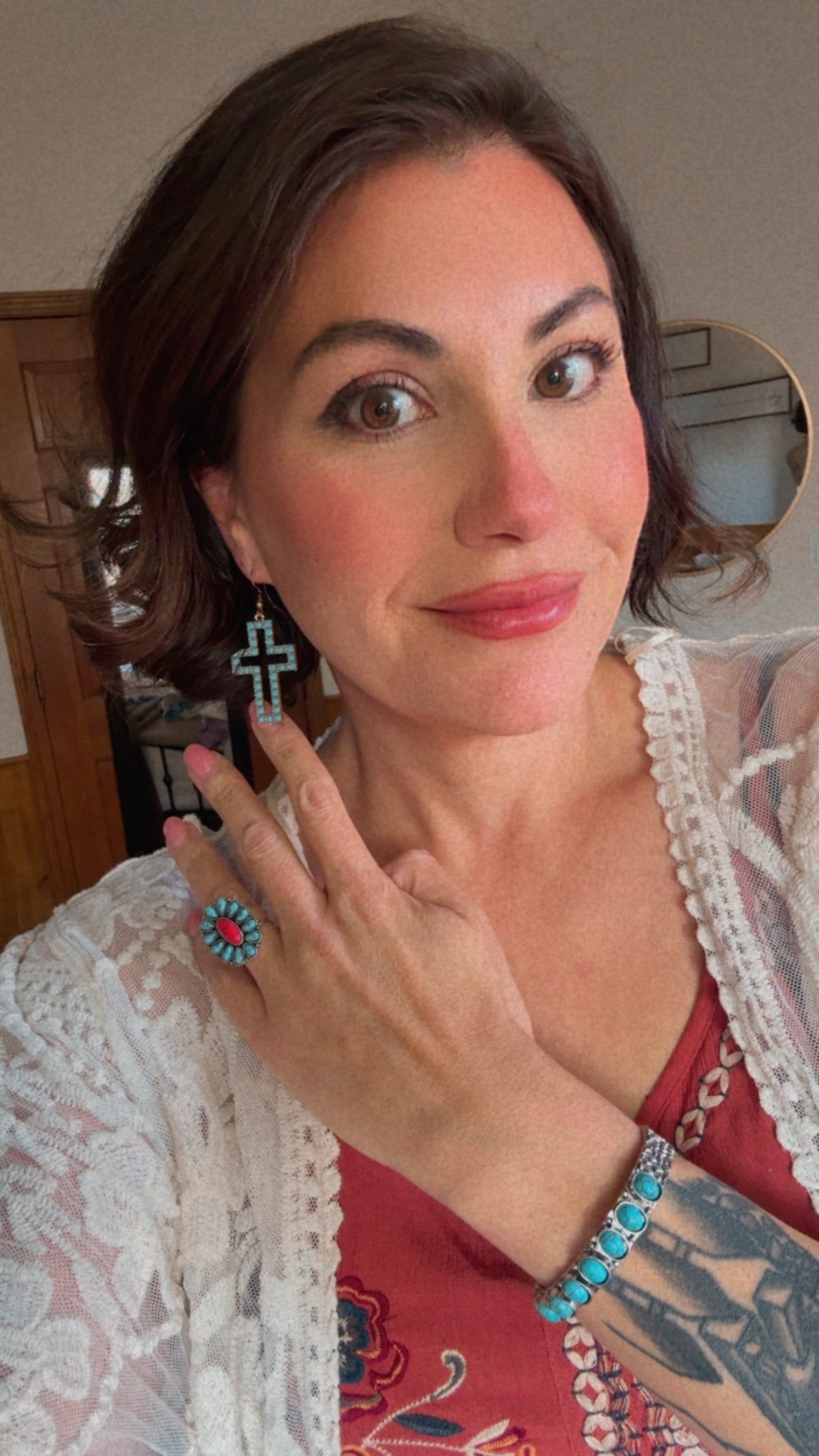 Photograph of a smiling woman indoors wearing a white lace cardigan over a red patterned top, accessorized with a turquoise cross earring, a coral and turquoise cluster ring, and a unique turquoise cuff bracelet