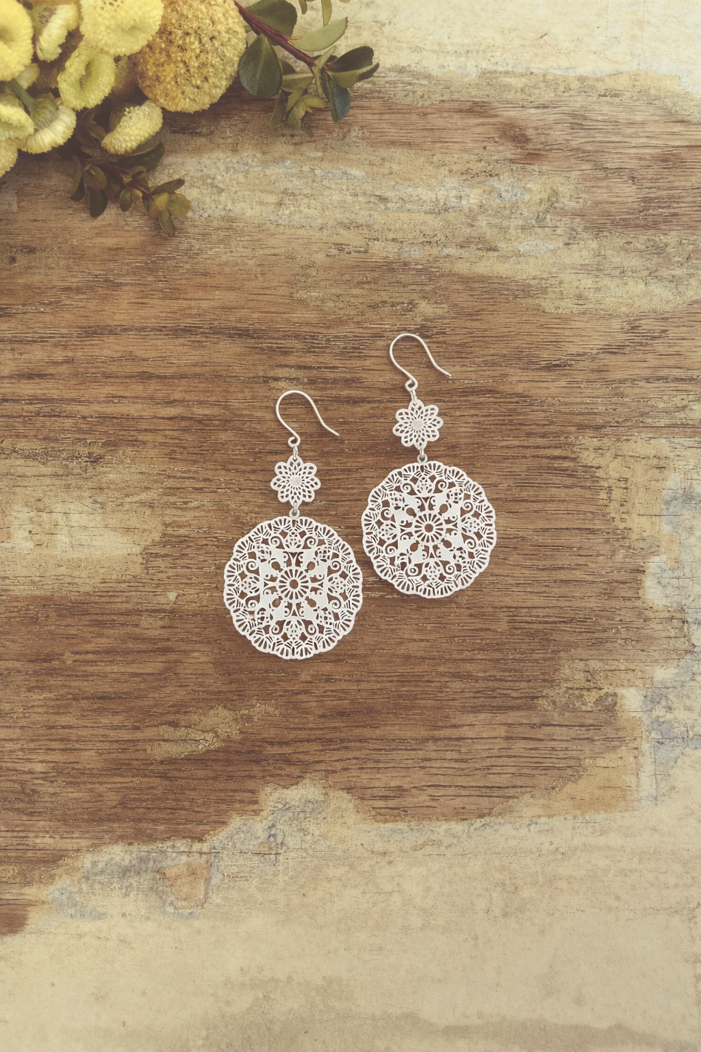Photograph of a pair of ornate silver filigree drop earrings with delicate floral designs resting on a weathered wooden table accented by vibrant yellow globe flowers