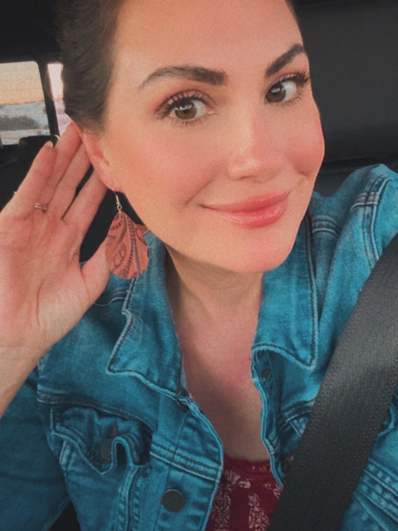 Digital photograph of a smiling woman in a car wearing a blue denim jacket and distinctive copper teardrop earrings with an embossed pattern, her left hand lightly touching her ear, with a glimpse of a red patterned top