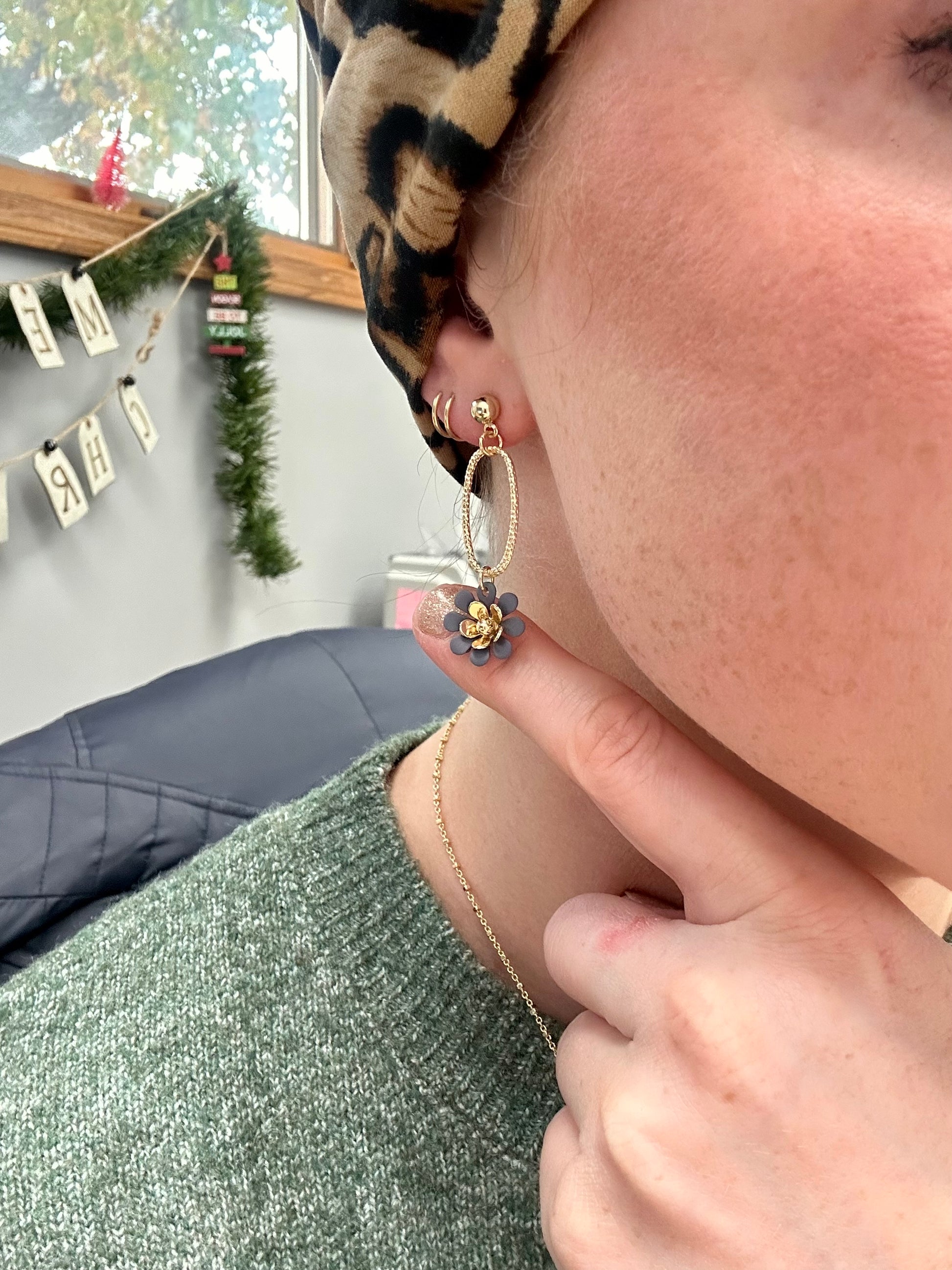 Close-up of a woman's ear wearing a gold dangle earring with a grey and gold flower charm, complemented by a leopard print head covering and green knit sweater, with a festive background featuring Christmas garland and a red bottle brush tree