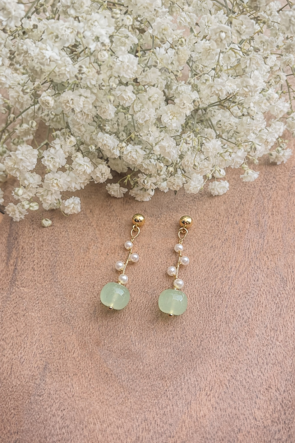 A photograph showcasing a pair of elegant gold-toned dangle earrings with faceted light green gemstone beads and delicate white pearls against a light brown wooden background adorned with fluffy white baby's breath flowers