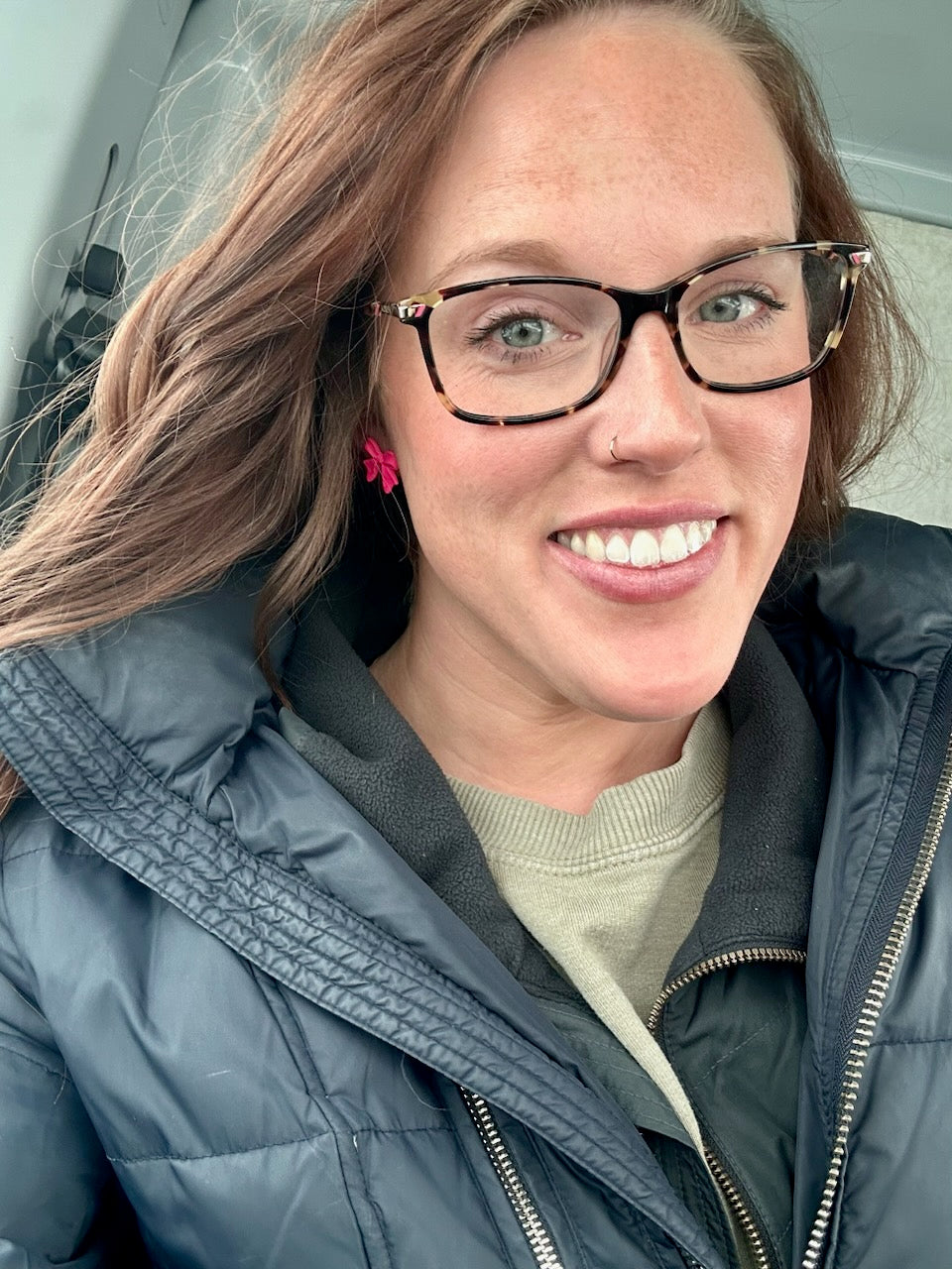 A close-up photograph of a smiling woman with reddish-brown hair, greenish-blue eyes, and a nose piercing, wearing tortoiseshell glasses, bright pink bow earrings, and a dark blue puffer jacket over a light green sweater