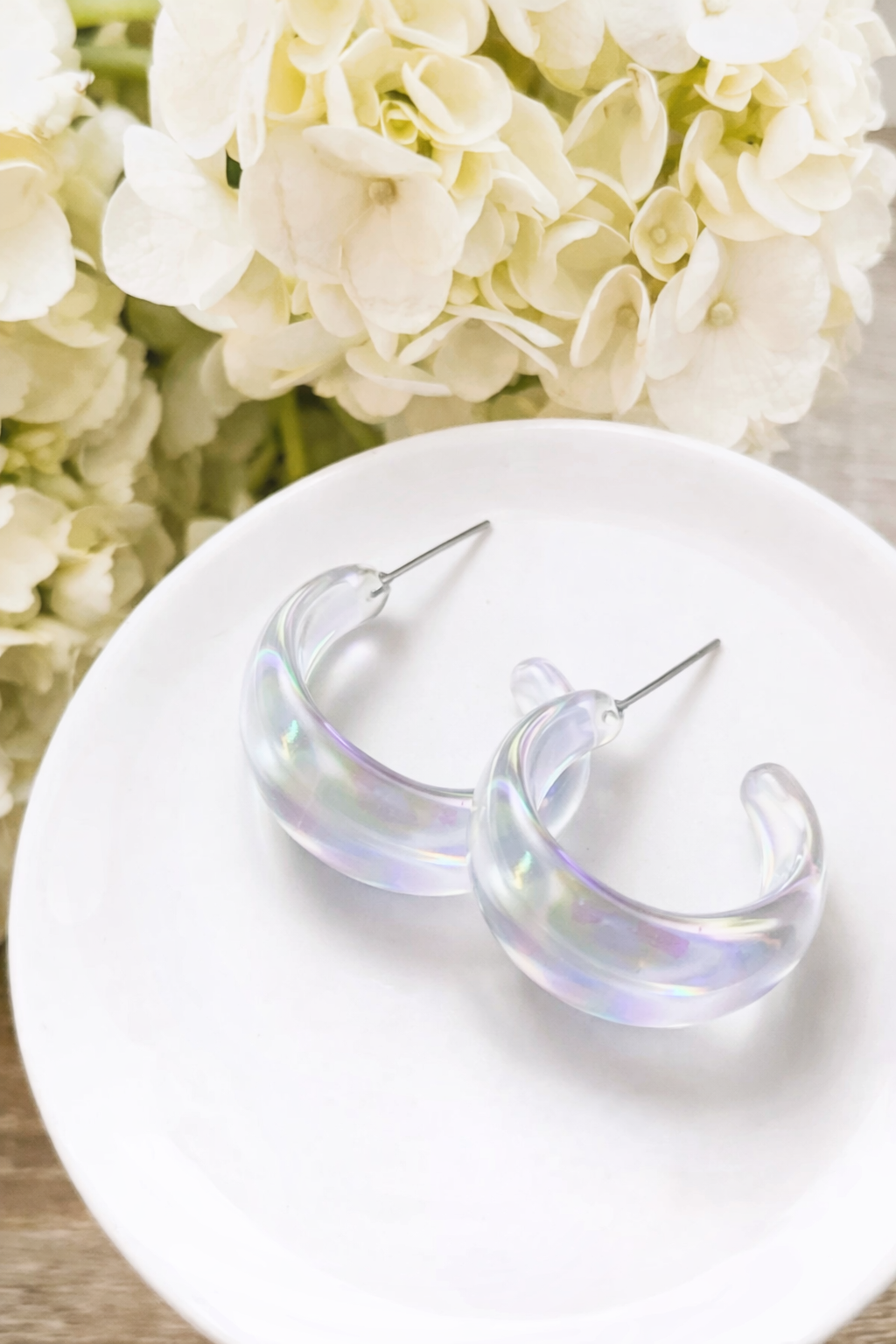Iridescent acrylic hoop earrings with silver-toned posts displayed on a pristine white ceramic plate against a soft-focus background of fluffy white hydrangeas