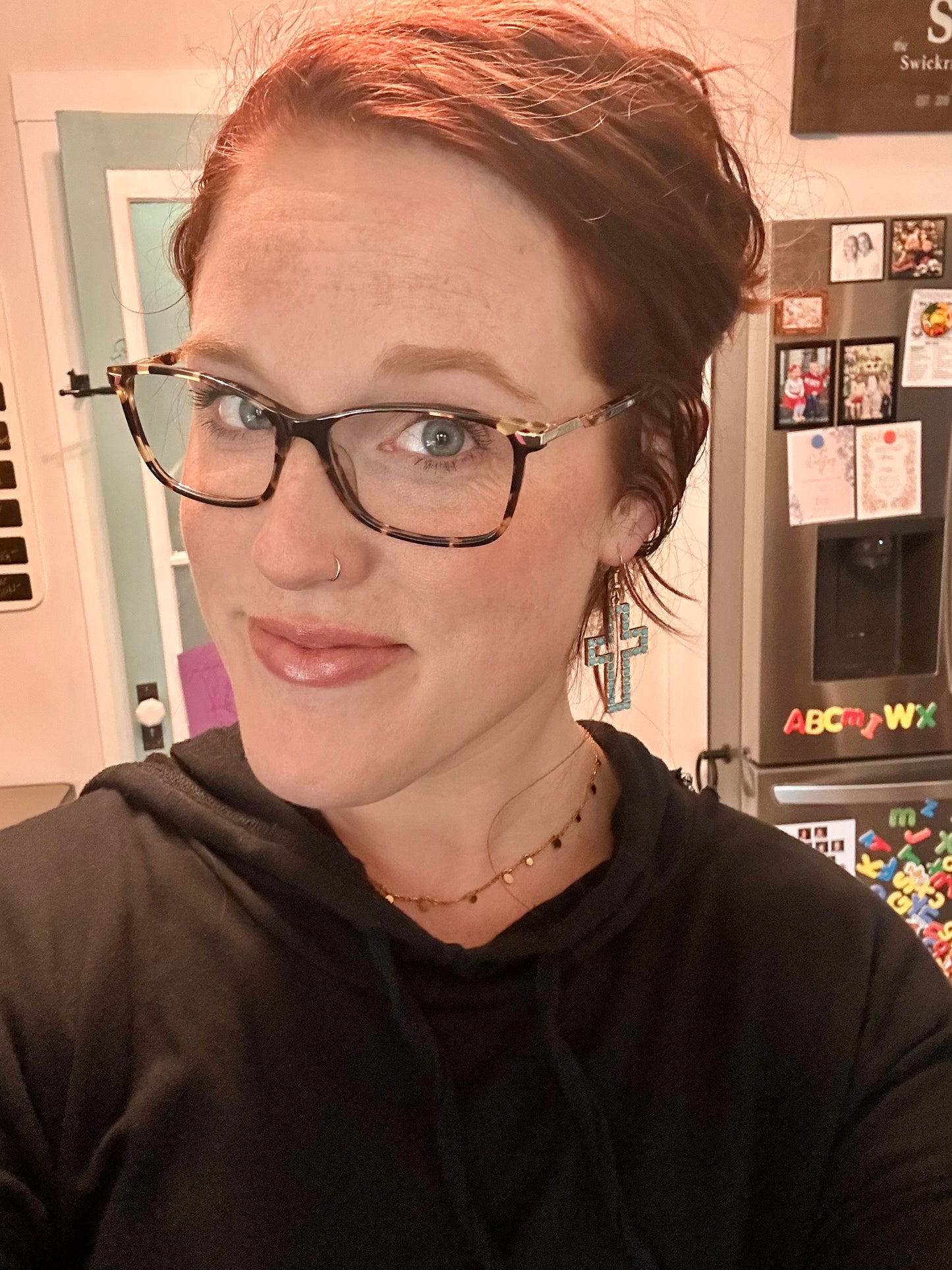 A smiling woman with reddish-brown hair, tortoiseshell glasses, a delicate gold nose ring, and turquoise cross earrings poses in a home kitchen in front of a refrigerator adorned with colorful alphabet magnets