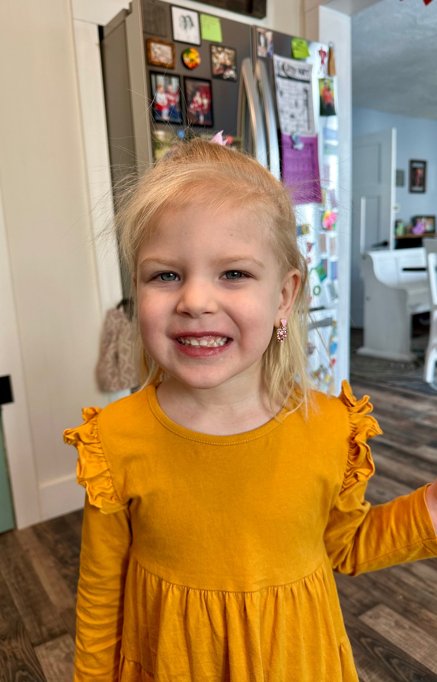 Photography of a smiling blonde girl in a mustard yellow ruffled dress indoors, featuring a pink heart earring and a pink hair clip