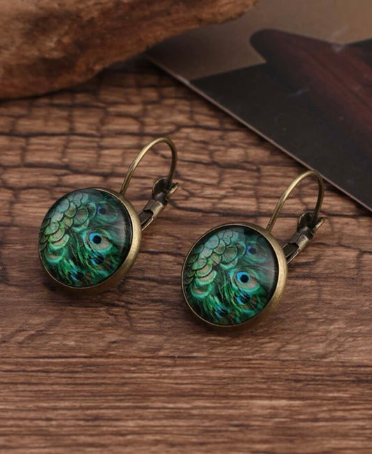 A pair of antique bronze lever-back earrings featuring a vibrant green and blue peacock feather cabochon design resting on a rustic wooden surface