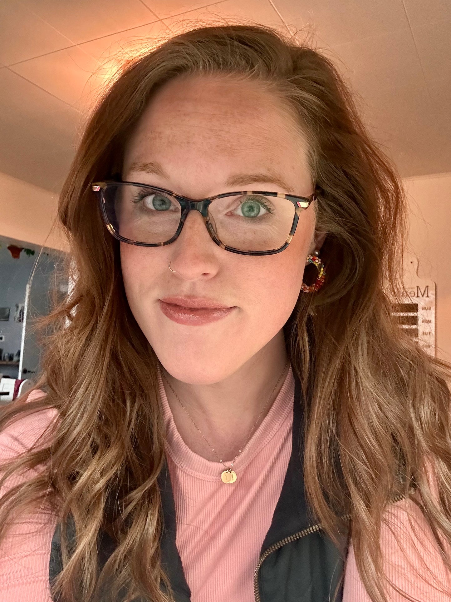 Digital photograph of a woman's indoor selfie featuring her freckled face, bright green eyes behind tortoise-shell glasses, and a vibrant beaded hoop earring