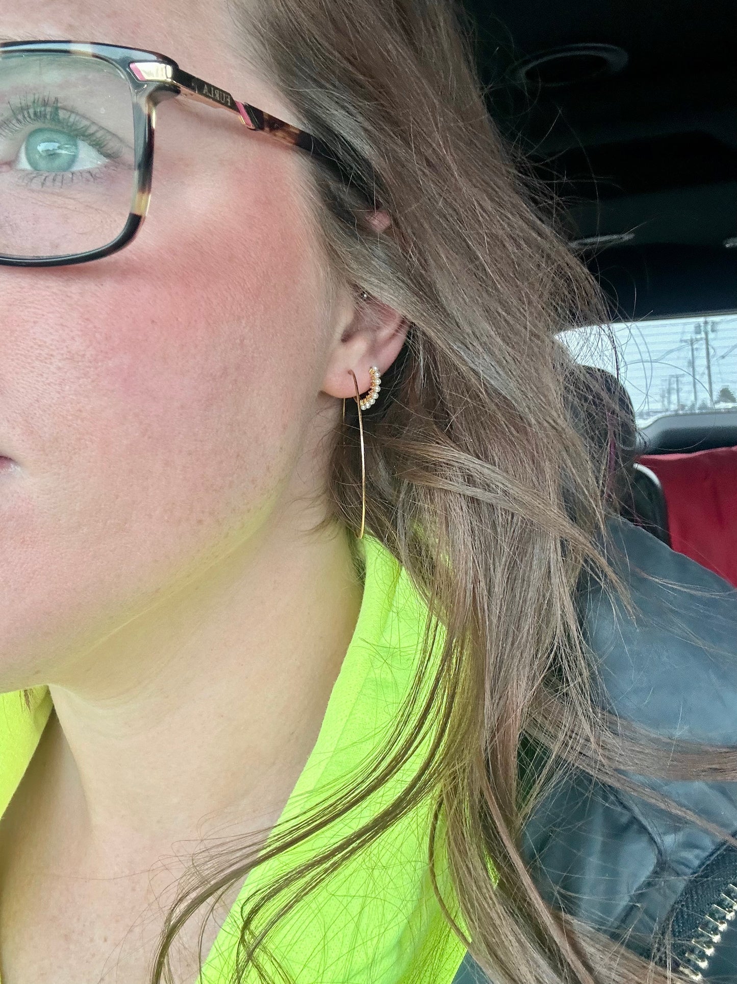 Photography of a woman's right profile inside a vehicle, featuring a light green eye behind tortoiseshell glasses, a delicate gold and pearl drop earring, and a vibrant neon yellow top peeking from under her long brown hair