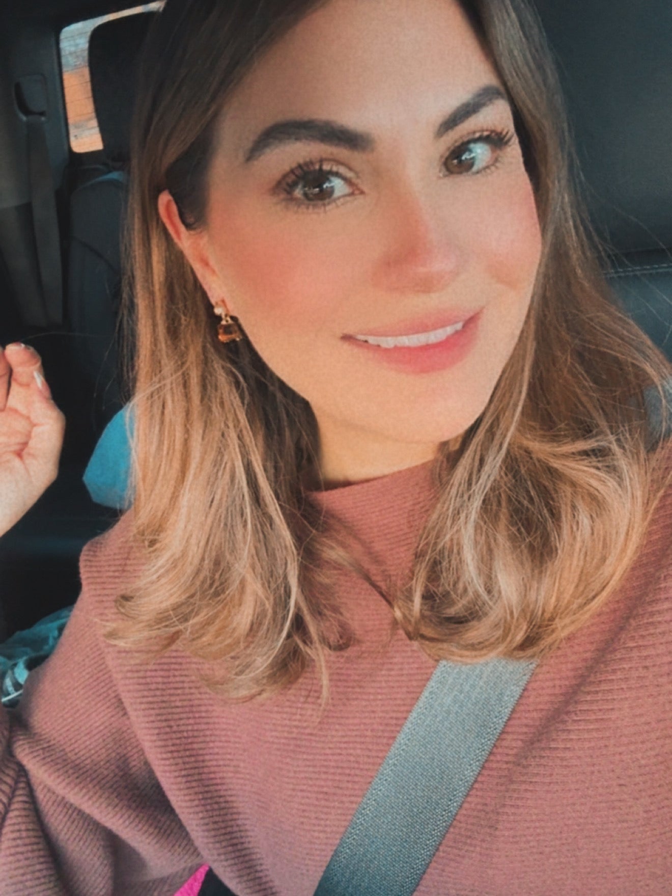 Photo of a smiling woman with light brown hair wearing a pink ribbed sweater and a safety belt in a car, featuring a small amber-colored dangling earring
