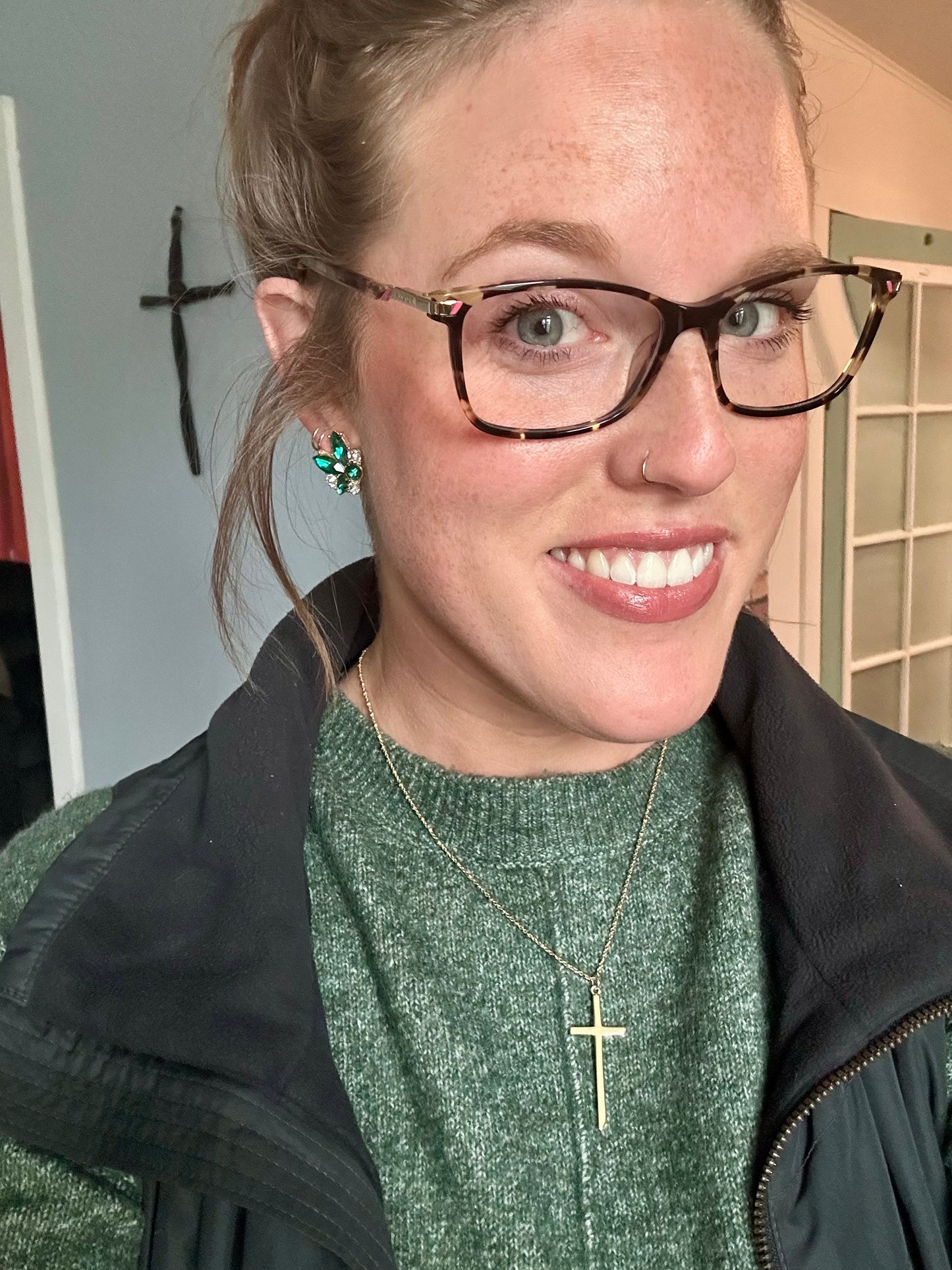 Smiling young woman with freckles wearing a green knit sweater, tortoiseshell glasses, sparkling emerald earrings, and a gold cross necklace, photographed indoors against a light wall with a subtle dark cross decoration