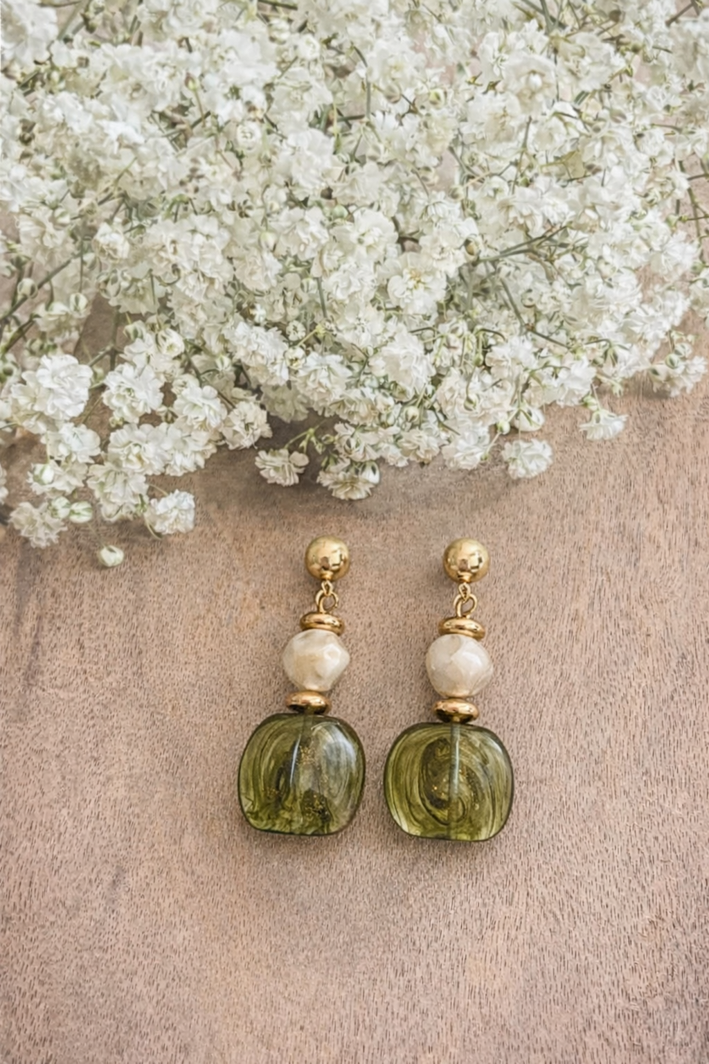 Photograph of a pair of decorative dangle earrings featuring gold posts, irregular milky white beads, and translucent olive green glass drops with swirling gold flecks, displayed on a light brown wooden surface below a cluster of delicate white baby's breath flowers