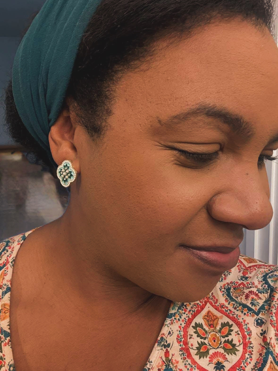 Photograph of a dark-skinned woman in a teal headscarf and a colorful paisley blouse featuring red, orange, and green patterns, wearing a prominent green and white jeweled clover earring