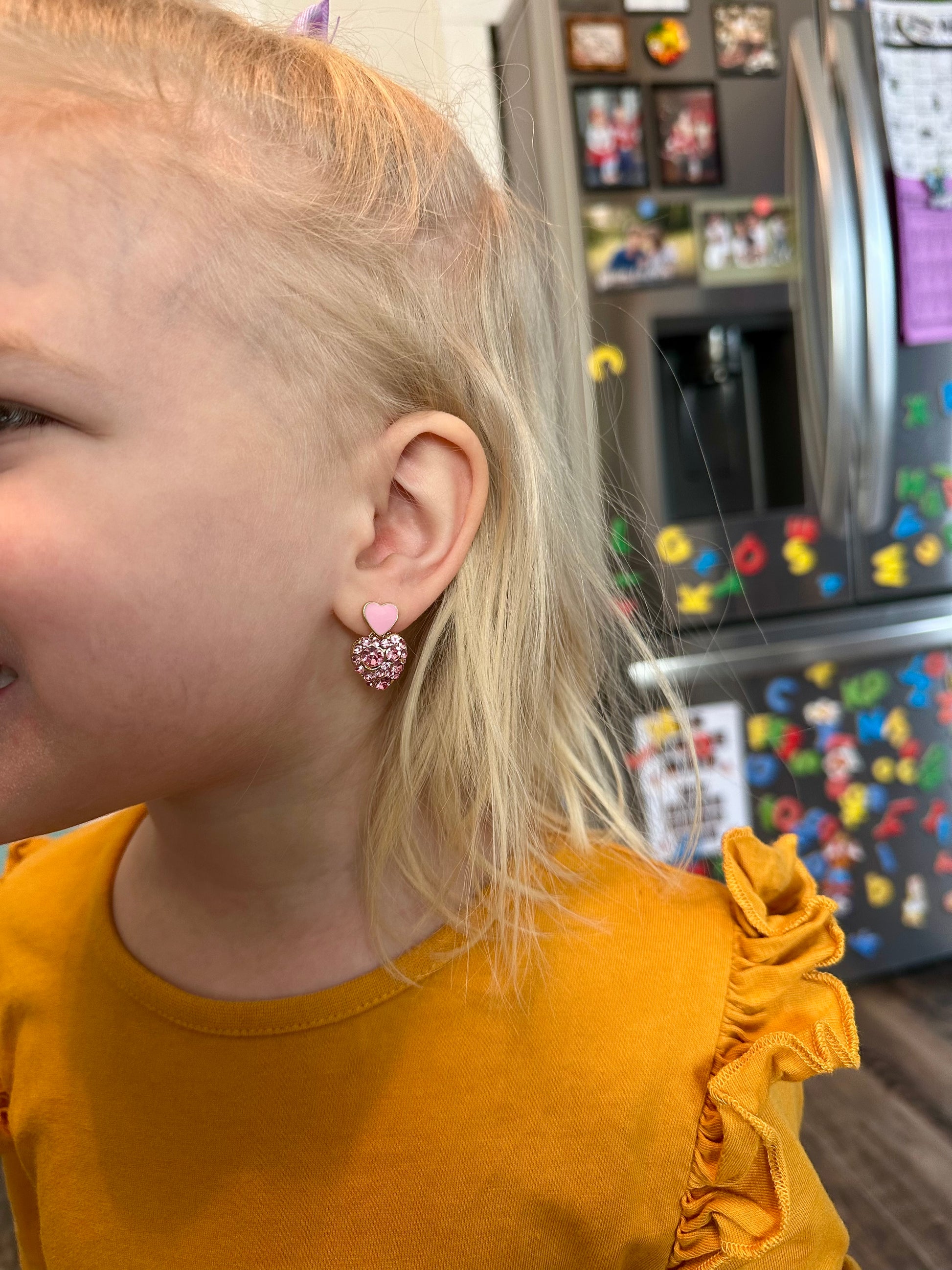 Close-up of a young blonde child's head and shoulder wearing a sparkly pink heart earring and a mustard yellow ruffled shirt with a blurred stainless steel refrigerator and colorful alphabet magnets in the background