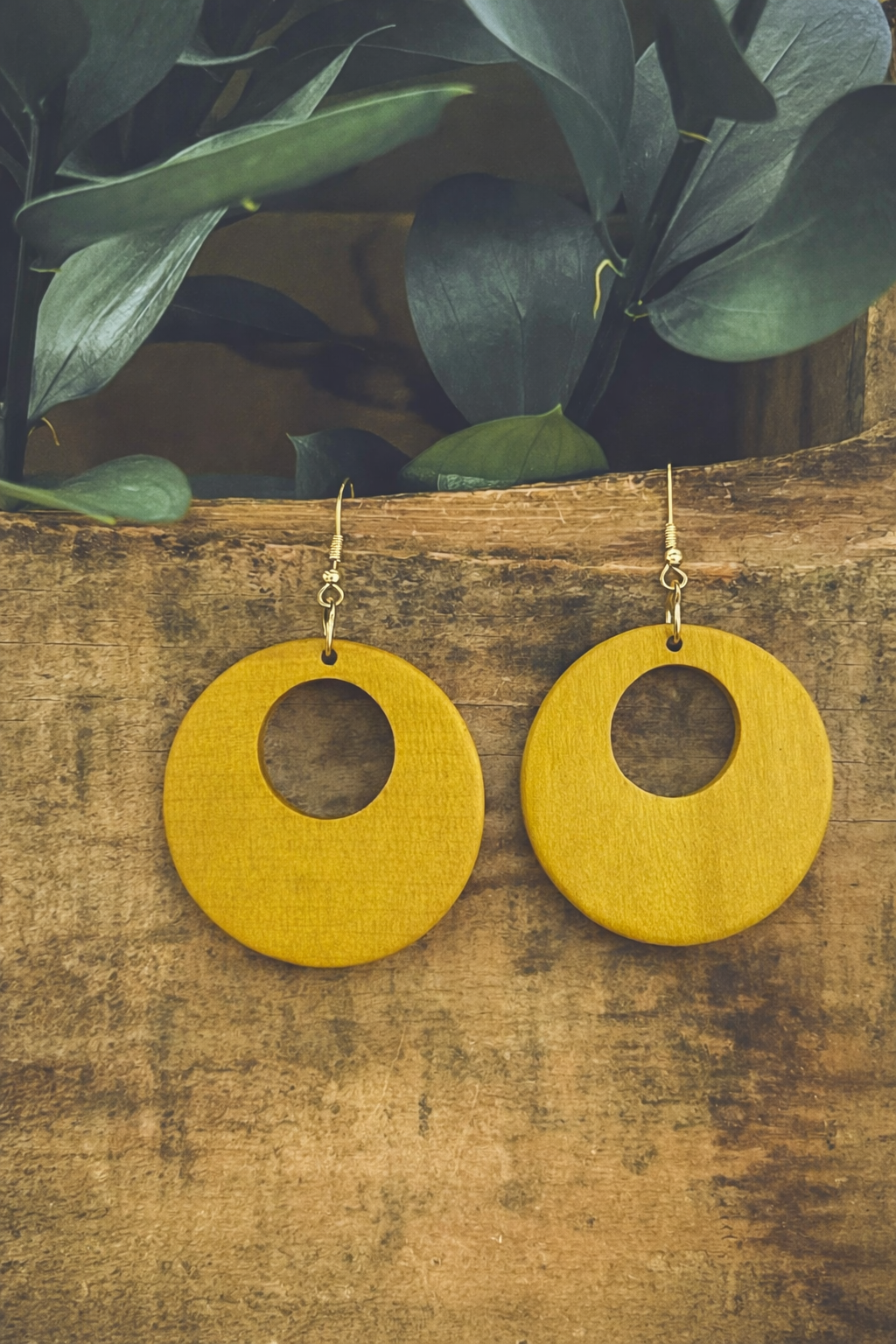 Product photo of two mustard yellow circular wooden earrings featuring central cutouts and gold-toned hooks, resting on a textured wooden plank with dark green leaves in the soft-focus background