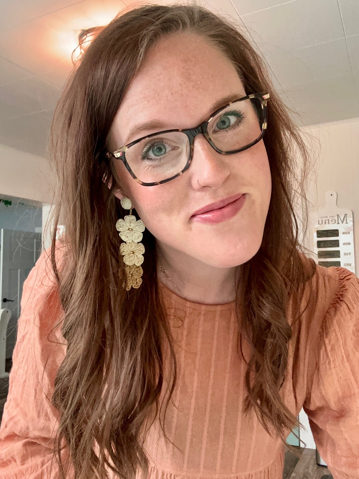 Selfie of a smiling woman with bright green eyes, tortoiseshell glasses, long brown hair revealing gray roots, wearing a peach textured blouse and ornate beige floral drop earrings in an indoor setting