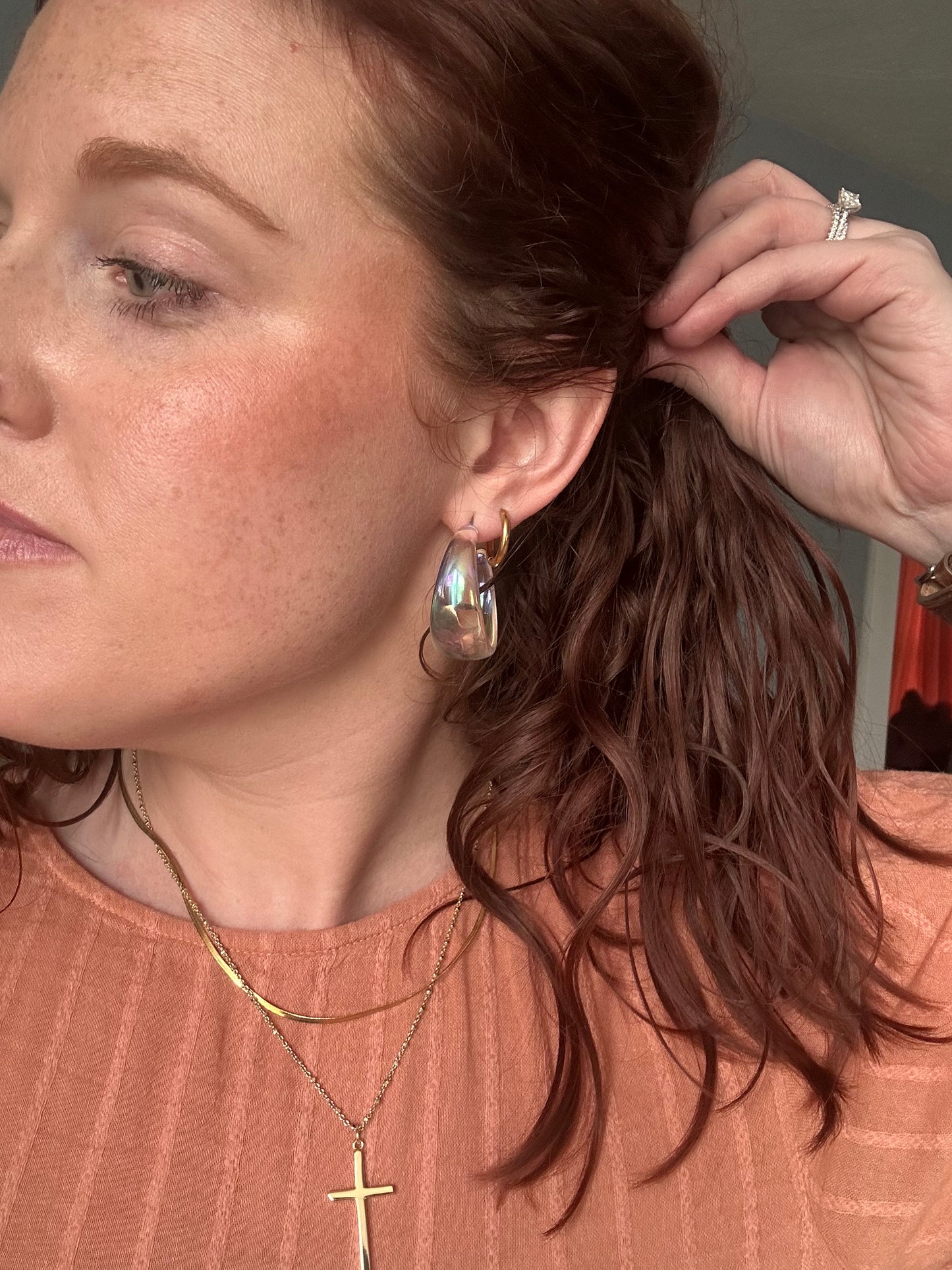 Photograph of a woman with reddish-brown wavy hair pulling it back, wearing an iridescent hoop earring, a layered gold cross necklace, and a sparkling diamond ring