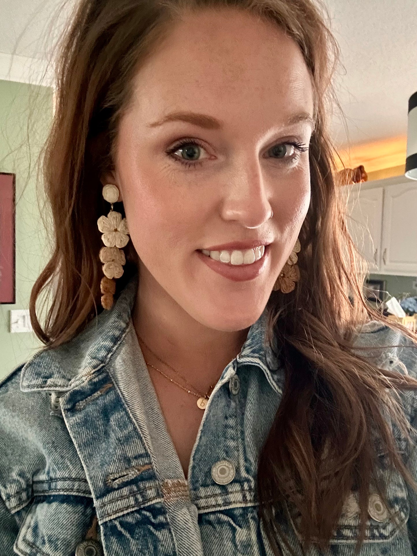 Photograph of a smiling woman with reddish-brown hair and green eyes, wearing a blue denim jacket, multi-tier floral earrings, a nose ring, and a gold 'b' initial necklace, in a home with a green wall and white cabinets