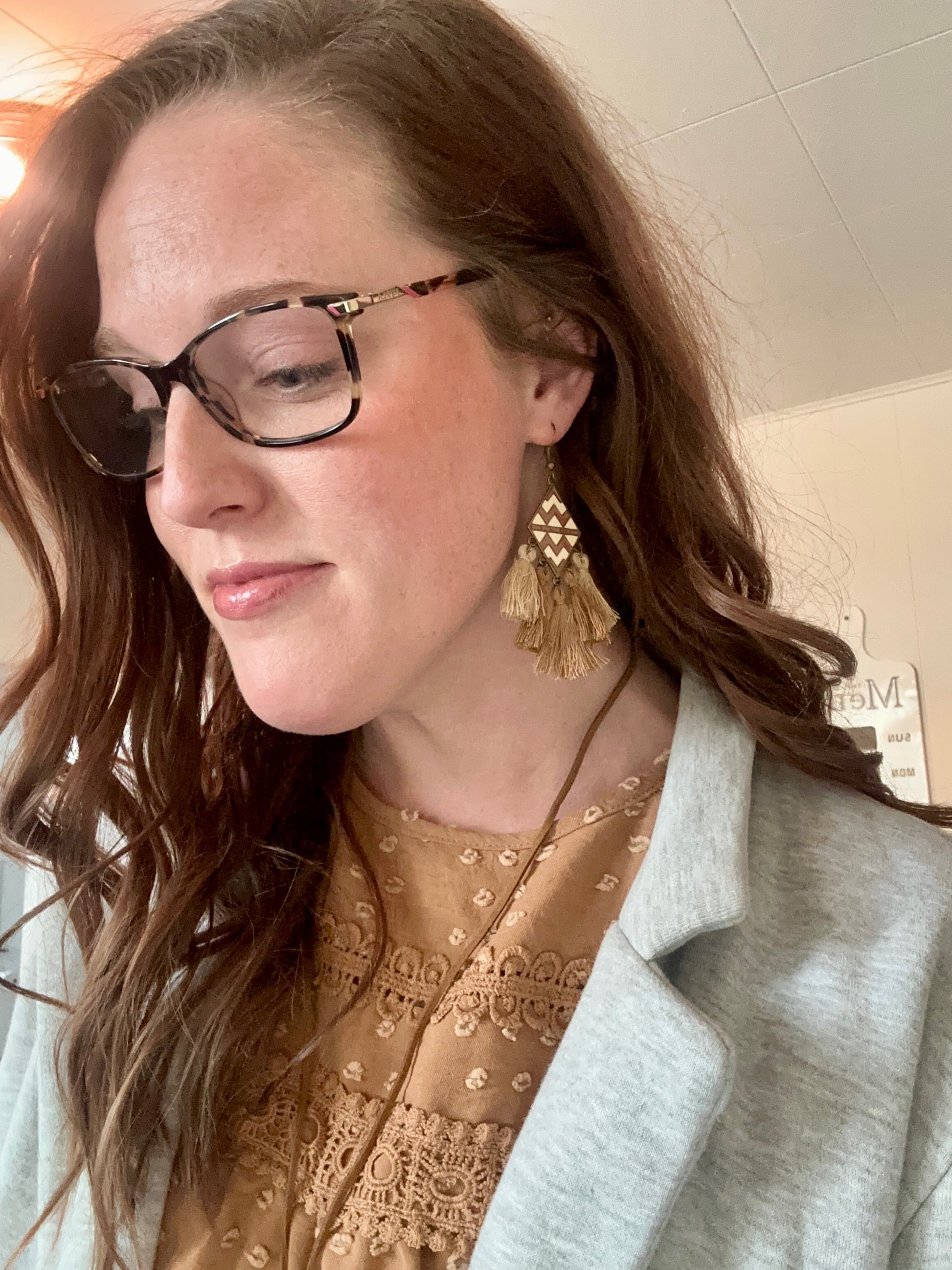 Close-up portrait of a woman wearing leopard print glasses, geometric gold and tan tassel earrings, and a tan top with lace and raised dot details