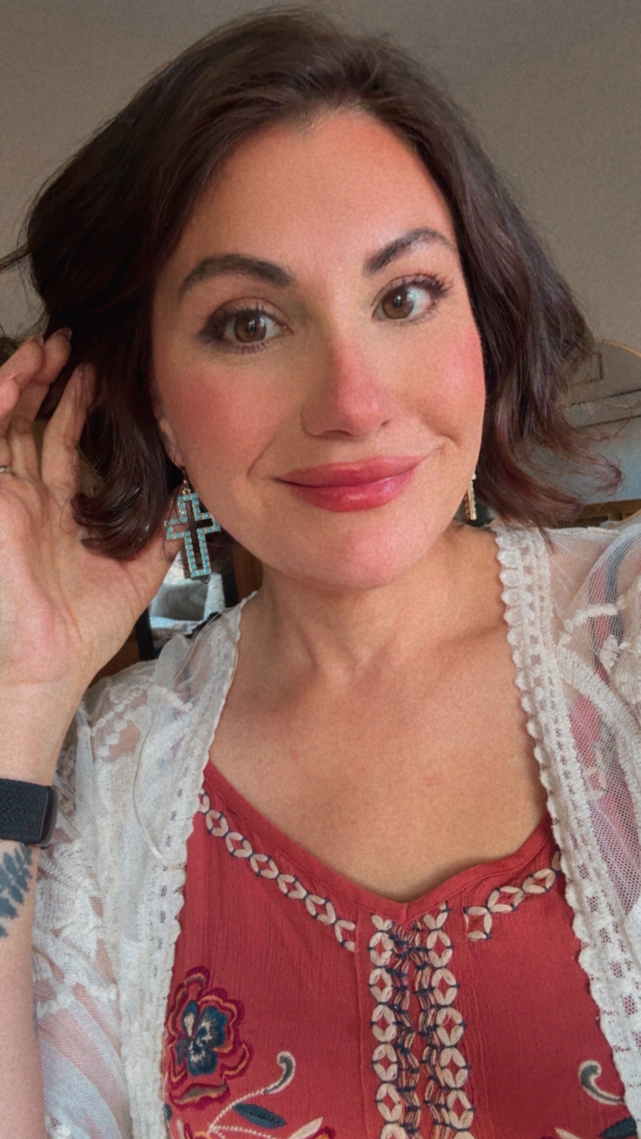 Smiling woman with dark brown hair wearing a burnt orange embroidered top, a white lace cardigan, and prominent turquoise cross earrings