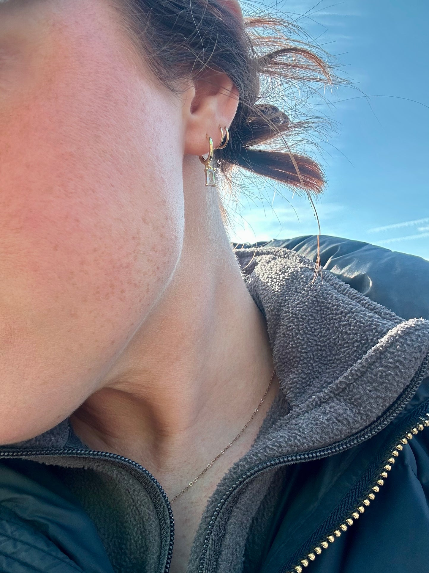 Photography of a person's profile with freckled skin and windswept brown hair, wearing a golden hoop earring with a clear rectangular crystal drop, a thin gold chain necklace, and a dark grey fleece jacket against a bright blue sky