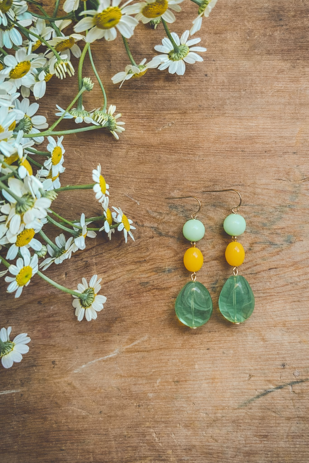 Artisanal dangle earrings with mint green, sunny yellow, and clear green teardrop beads rest on a rustic wooden surface next to a cluster of white daisies with yellow centers