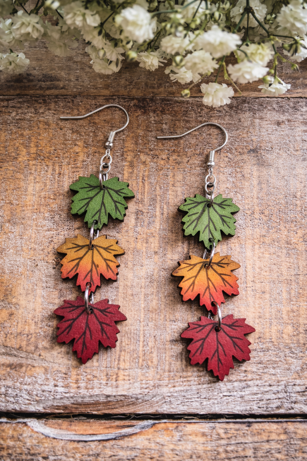 A pair of dangling earrings featuring wooden maple leaf shapes in a gradient of green, orange, and red with etched veins and silver hooks, resting on a rustic wooden surface under blurred white baby's breath flowers