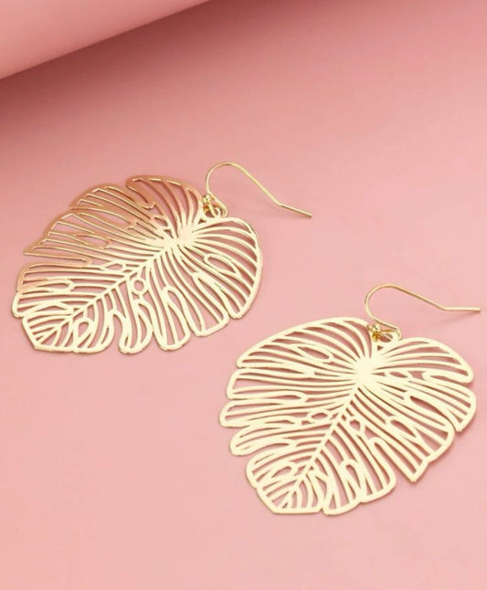 A pair of gold monstera leaf earrings featuring an intricate filigree design and hook closures on a soft pink background