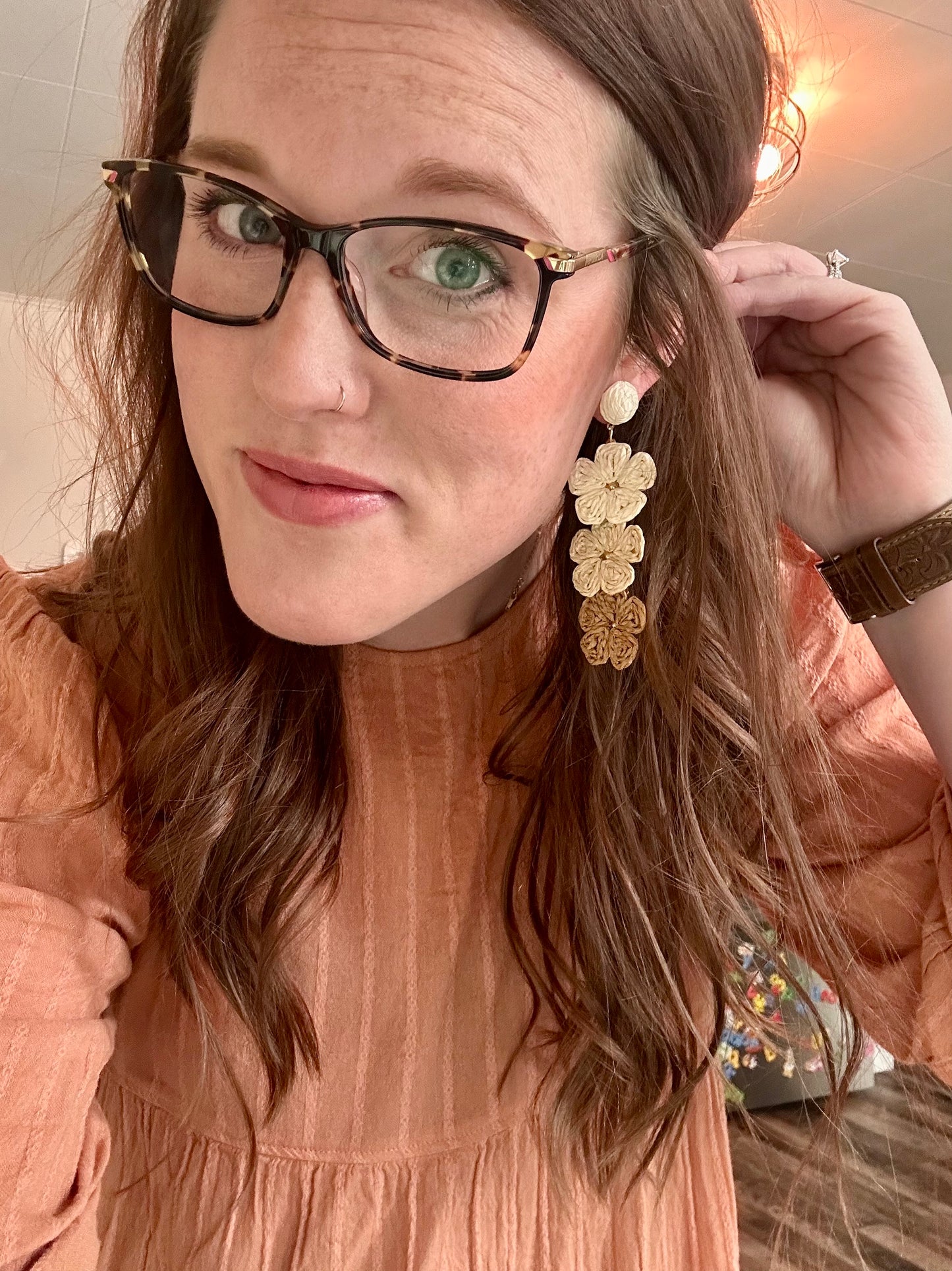 A smiling woman with green eyes, long brown hair, and tortoiseshell glasses featuring a gold accent, wearing a peach textured dress, a copper nose ring, and long stacked raffia flower earrings