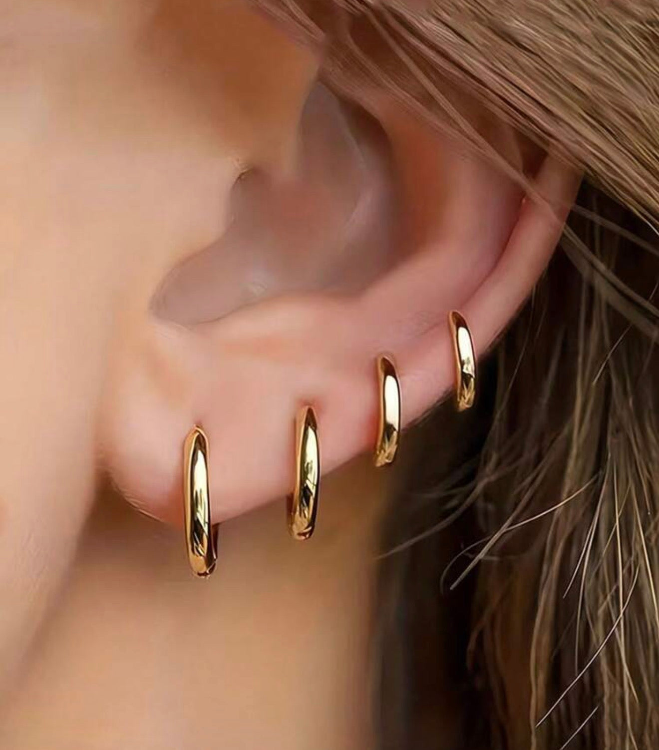 A pierced earlobe is adorned with four graduated shiny gold hoop earrings against a background of soft brown hair