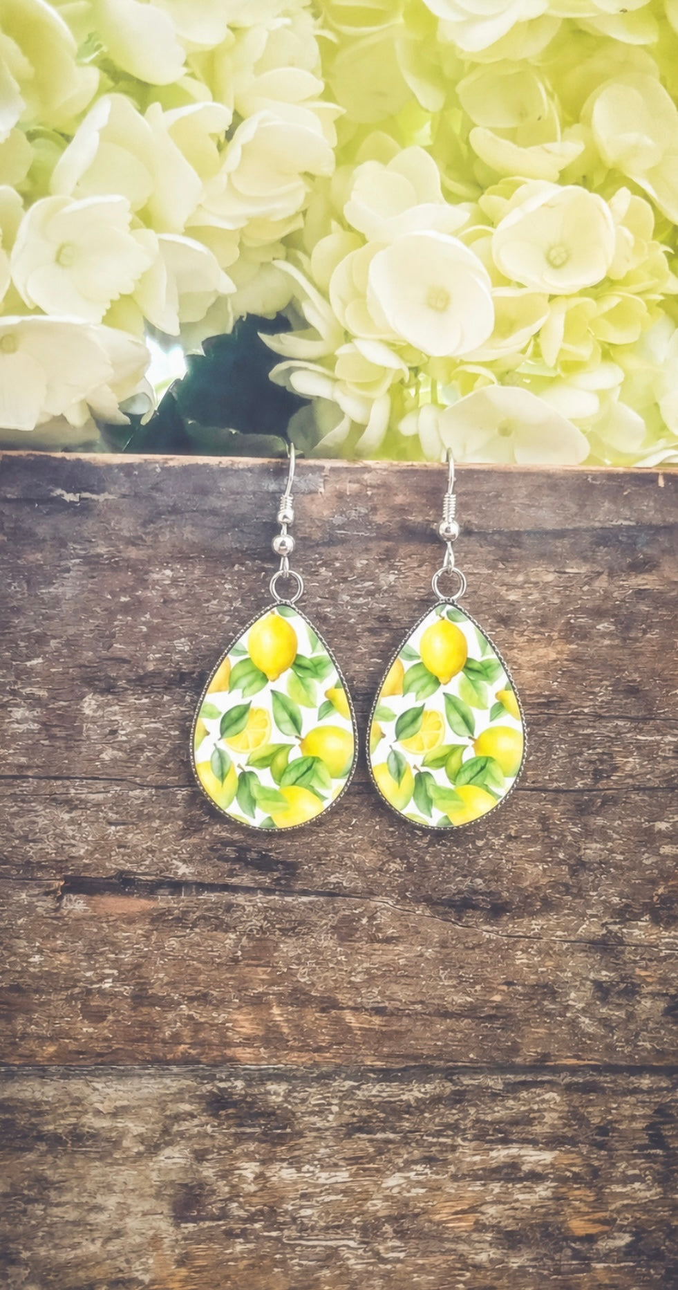 Handcrafted silver teardrop earrings with a vibrant lemon and green leaf pattern displayed on rustic wood planks against a backdrop of soft white hydrangeas