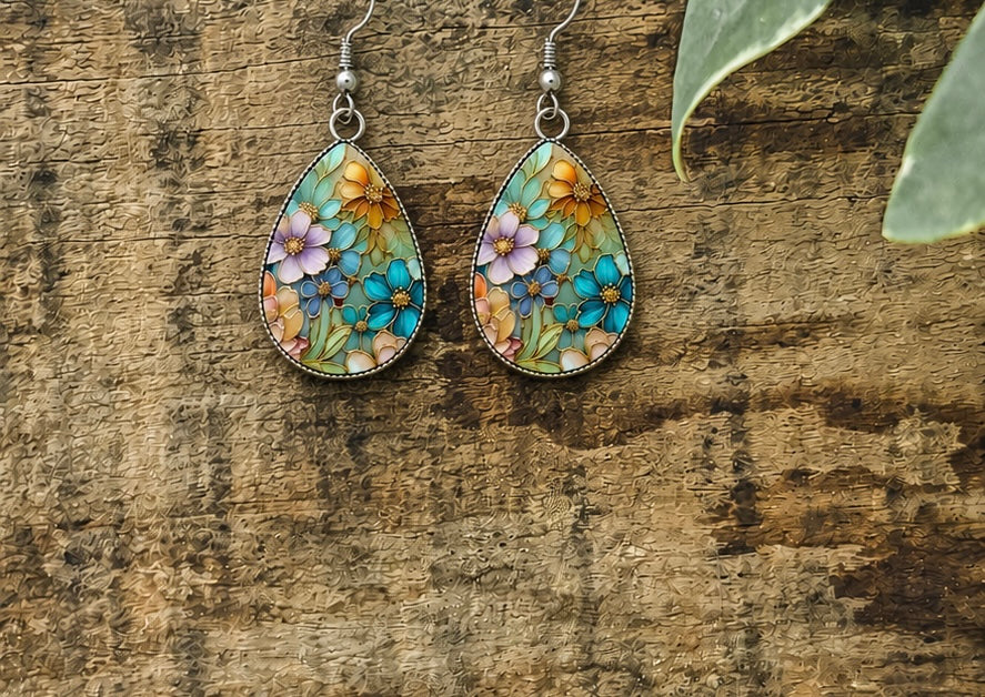 Handcrafted teardrop earrings with intricate vibrant purple blue and orange floral designs and green leaves displayed against a rustic weathered wooden backdrop