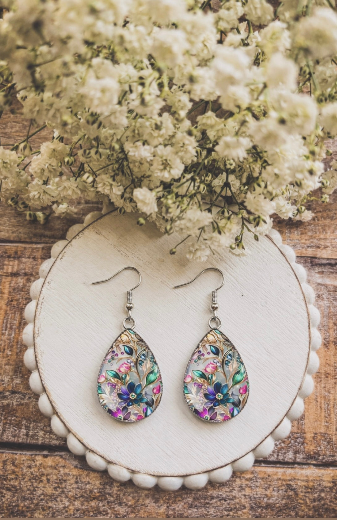 Handcrafted floral enamel teardrop earrings featuring vibrant purple pink teal and green designs presented on a white scalloped board over rustic wood with blurred white baby's breath flowers