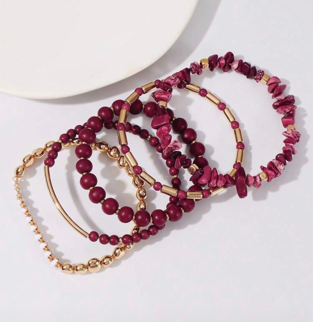 A layered set of bohemian bracelets featuring maroon round beads fuchsia stone chips gold cylindrical spacers and a delicate gold chain with white accents against a crisp white background