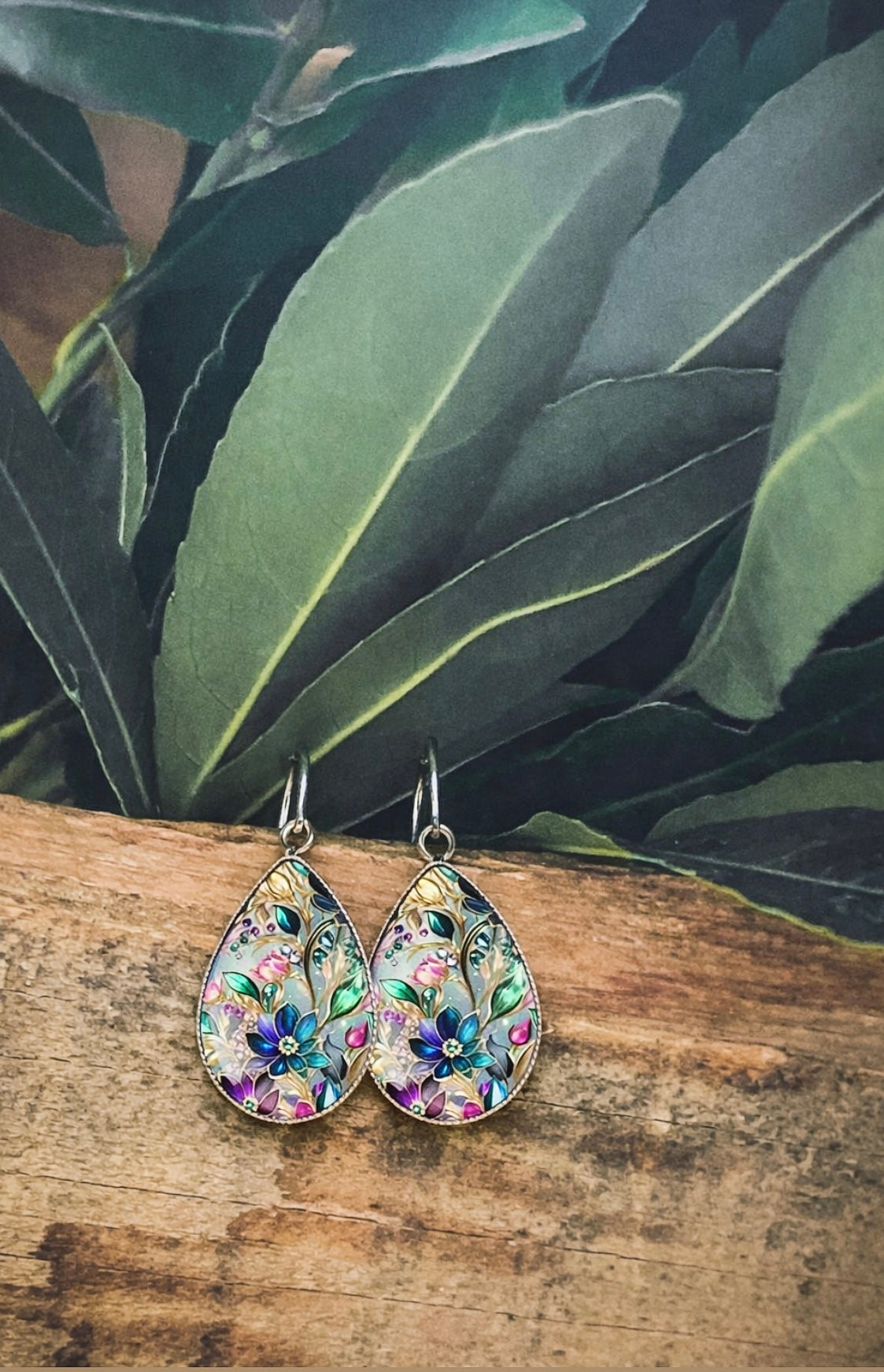 A pair of teardrop-shaped enamel earrings with vibrant floral patterns in teal, purple, pink, and gold, displayed on a rustic wooden surface against a backdrop of deep green leaves