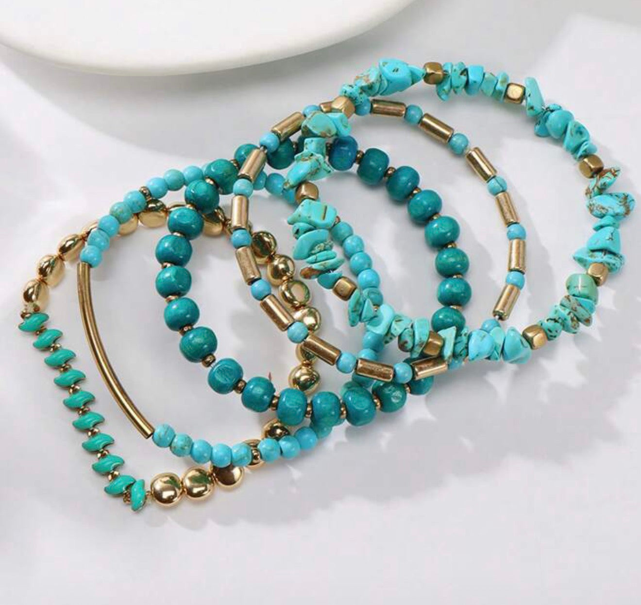 Photograph of a stack of five bohemian style bracelets on a white surface featuring vibrant blue-green turquoise beads in round and irregular chip shapes alongside gleaming gold-toned cylindrical and spherical elements