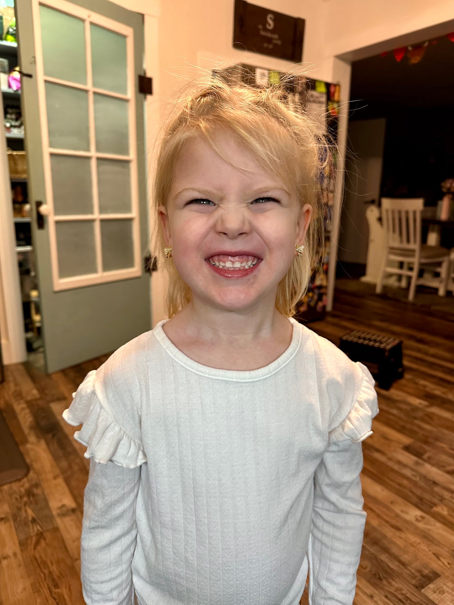 Photograph of a joyful blonde young girl with flyaway hair making a silly scrunch face and showing teeth wearing a white ruffled shirt and small bow earrings standing in a cozy home with a frosted glass pantry door and colorful string lanterns in the background