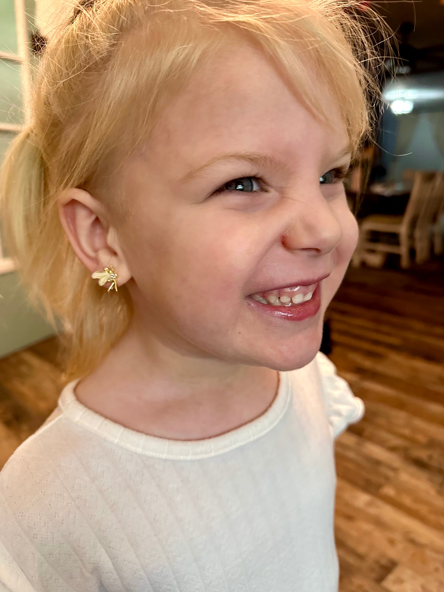 Photograph of a smiling young blonde girl with blue eyes and a gold bee earring wearing a white patterned shirt indoors