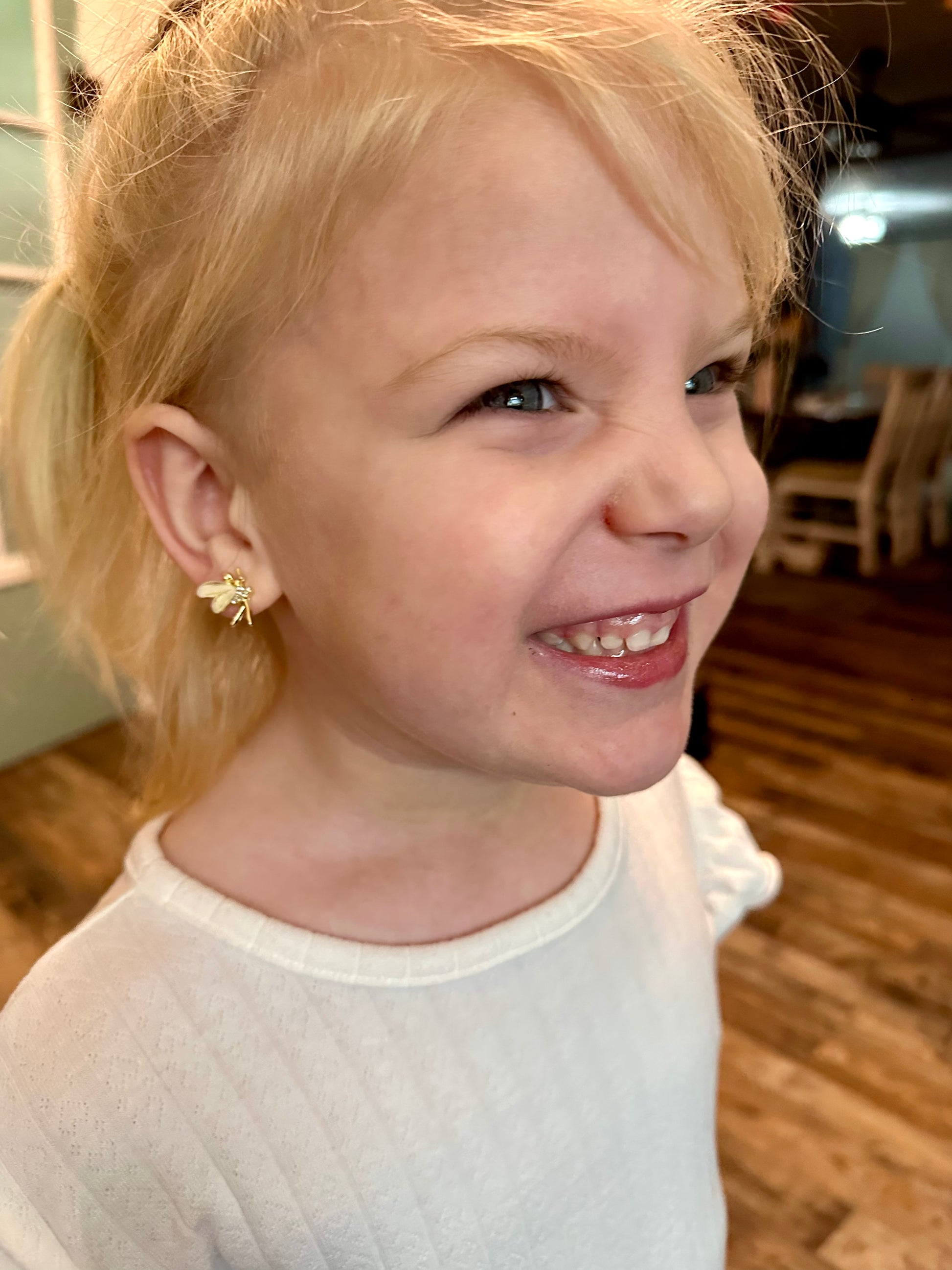 Photograph of a smiling young blonde girl with blue eyes and a gold bee earring wearing a white patterned shirt indoors