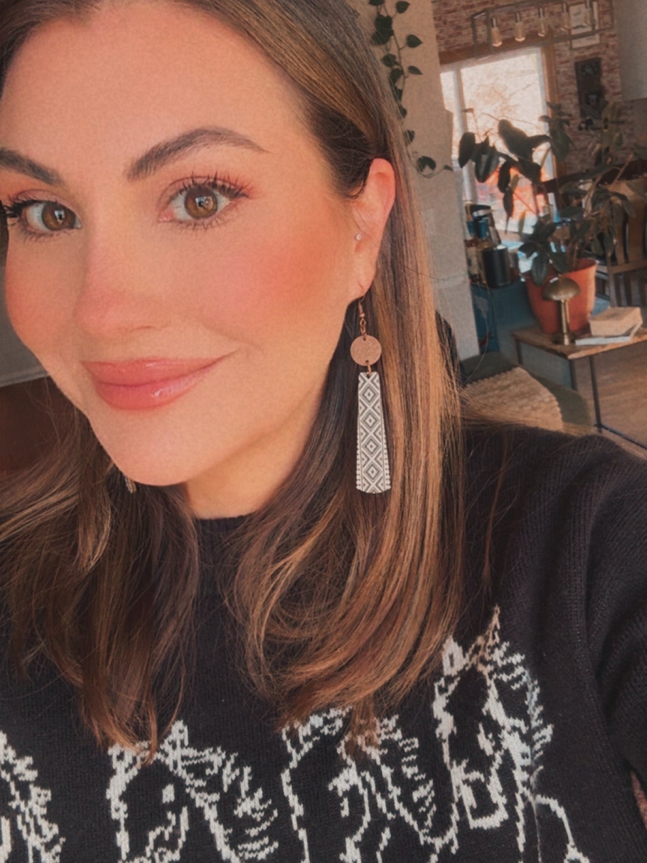 Photograph of an unidentified smiling woman with brown hair, rosy cheeks, and glossy lips, wearing a black sweater with a white geometric Fair Isle pattern and a long copper and white diamond-patterned dangle earring, in a sunlit home interior
