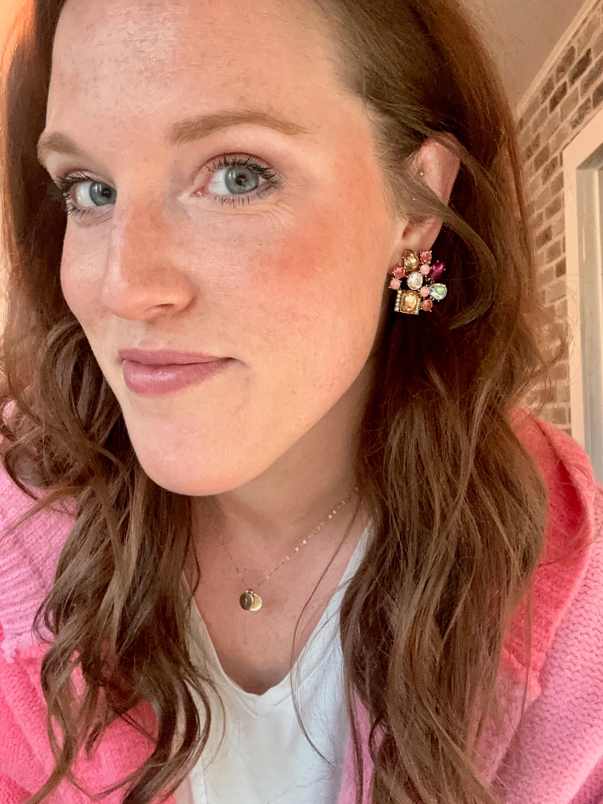 Photograph of a woman with reddish-brown hair and light blue eyes, wearing a bright pink knit sweater and a vibrant multicolored jeweled earring, indoors with a brick wall background