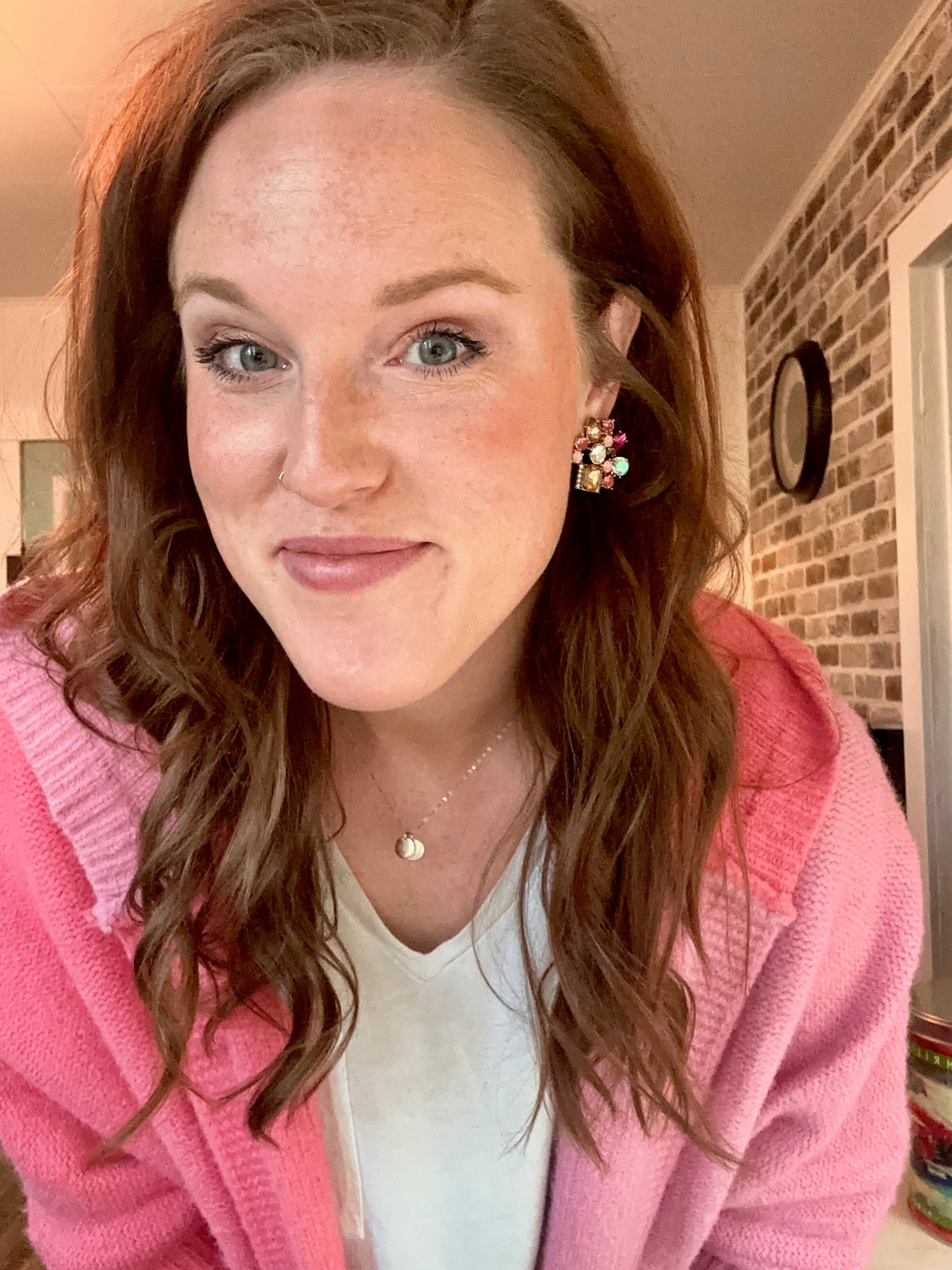 Digital photograph of a woman with long brown hair wearing a bright pink knitted cardigan, a colorful cluster earring, and a subtle nose ring, in front of a brick wall