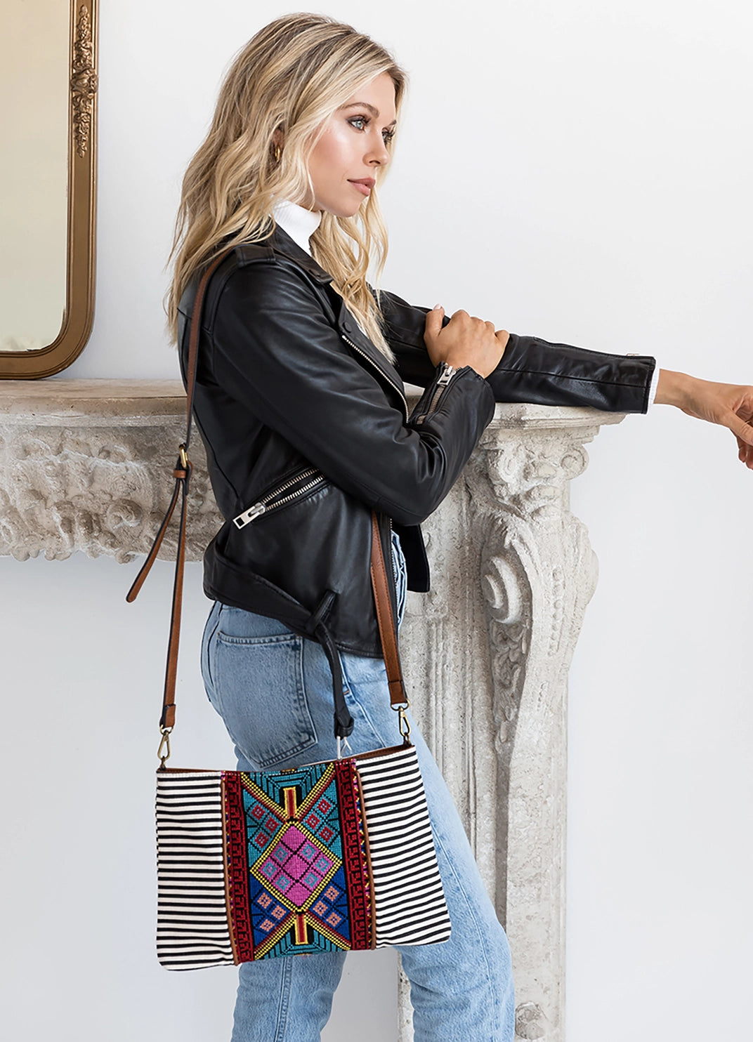 A blonde woman in a black leather jacket and blue jeans poses with a vibrant geometric and striped crossbody bag leaning on an ornate white stone fireplace mantel with a gold mirror in the background