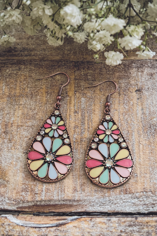 Photography of two ornate copper-tone teardrop dangle earrings featuring pastel enamel floral patterns in pink, yellow, blue, and mint green with small clear rhinestones, resting on a rustic wooden surface with blurred white baby's breath flowers above