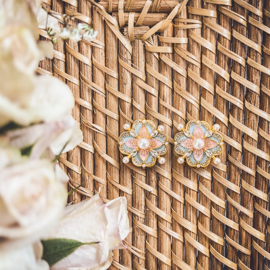 Ornate pastel pink and blue floral earrings with central pearls and intricate gold borders on a woven basket with blurred white flowers